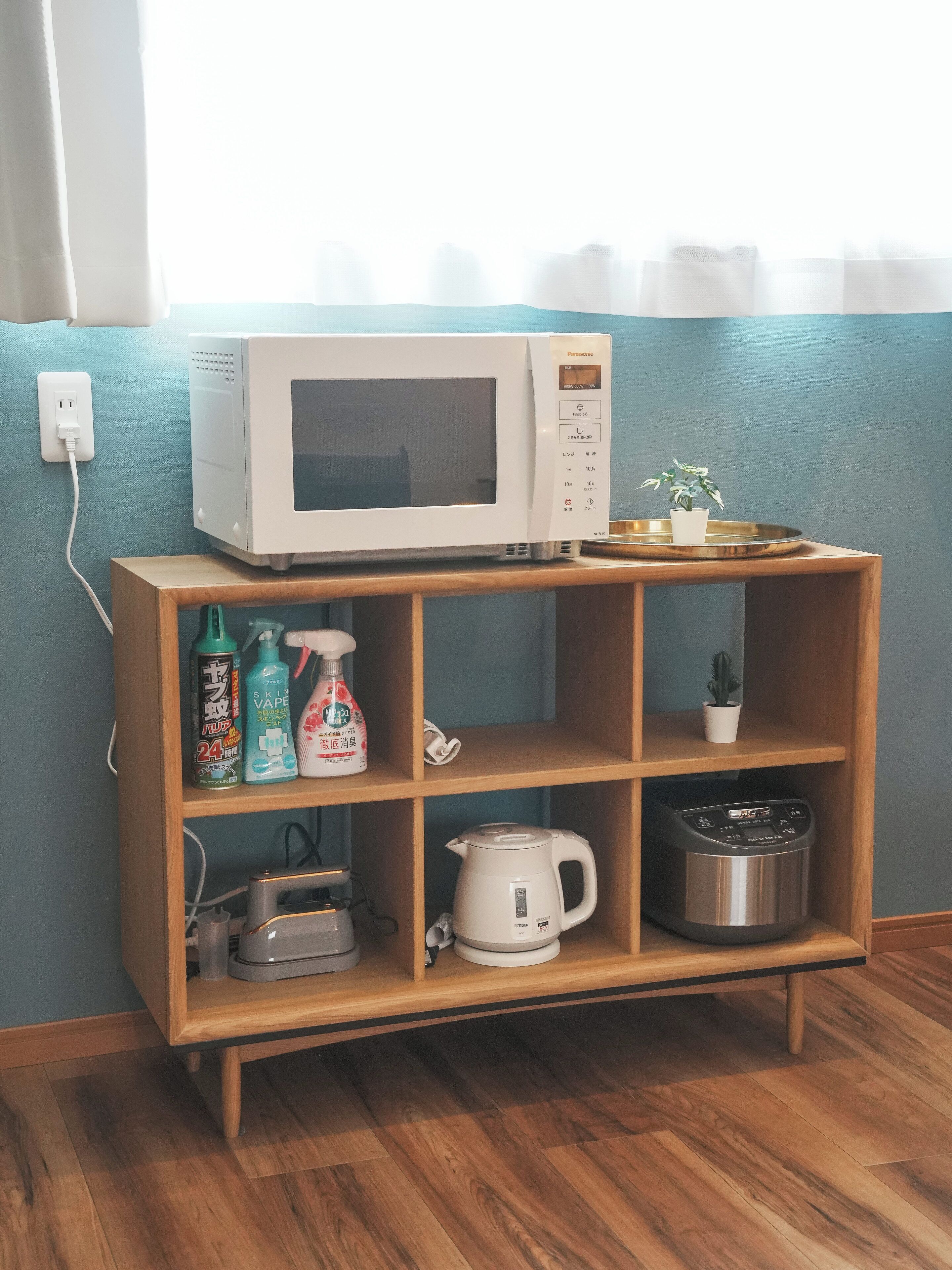 Un four à micro-ondes posé sur une étagère en bois, avec une plante en pot, une bouilloire et des produits de nettoyage.