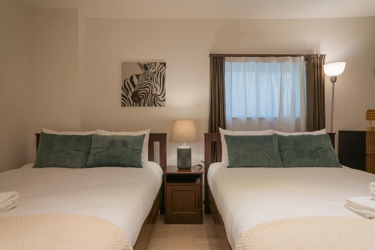 A hotel room with two beds, a nightstand with a lamp, a zebra artwork on the wall, and a window with curtains.