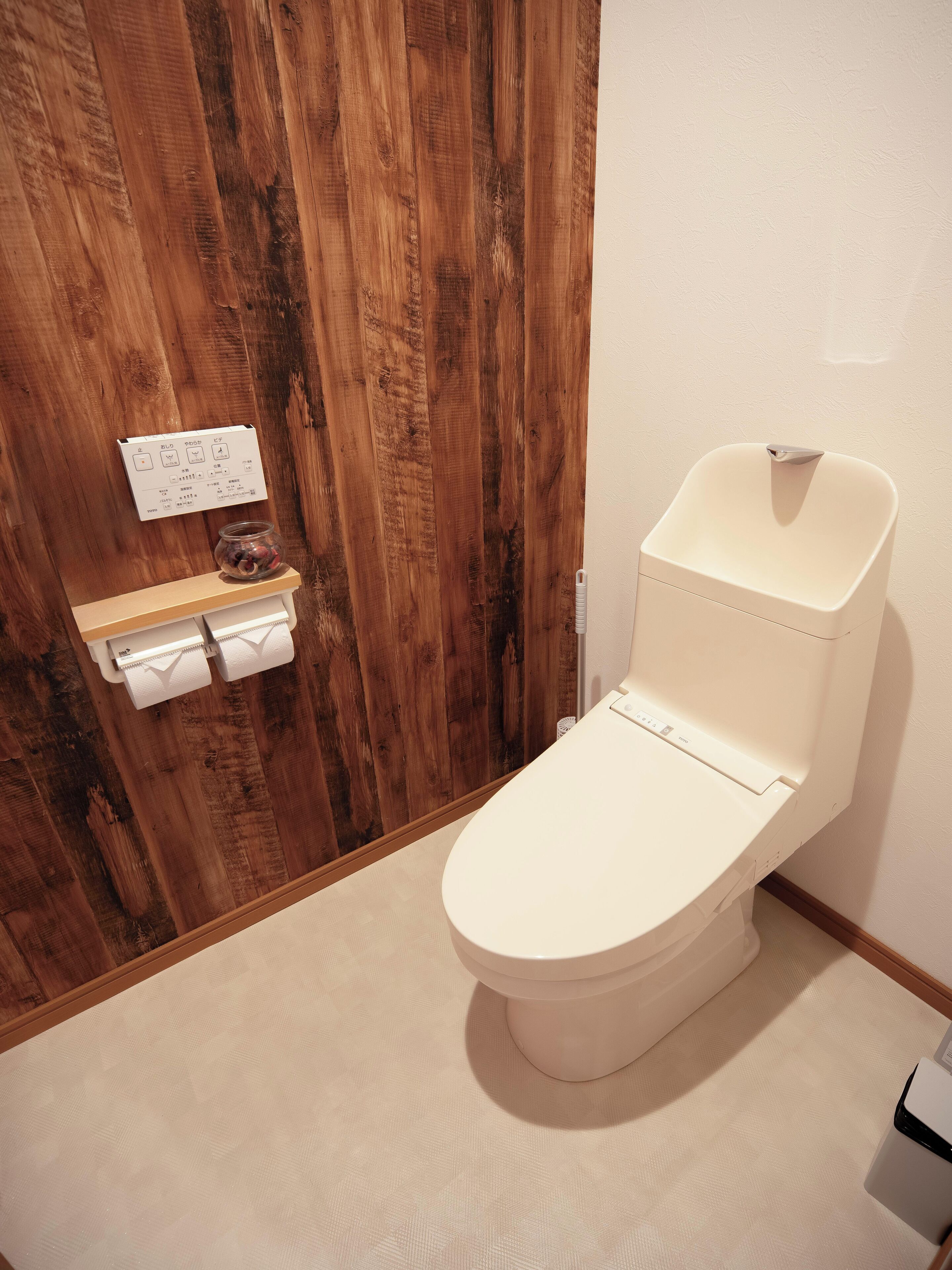 Un WC con sedile e serbatoio bianchi, una porta in legno con una lavagna e un piccolo ripiano con un vasetto e carta igienica.