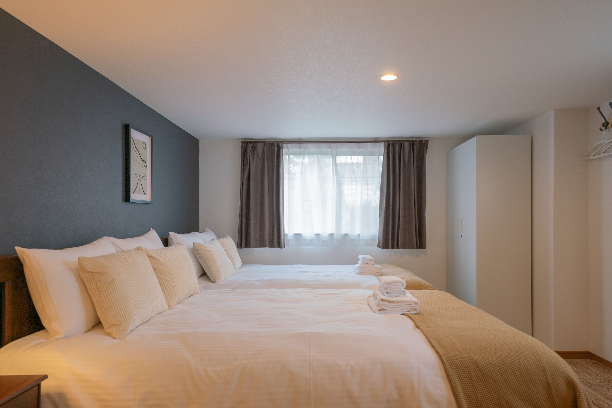 A hotel room with two beds, a large window with curtains, a wardrobe, and a framed picture on the wall.