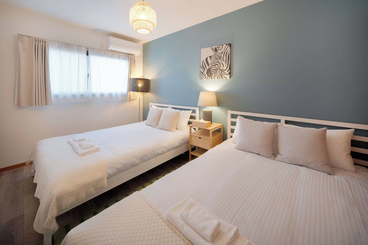 A hotel room with two beds, a nightstand with a lamp, a zebra artwork on the wall, and a window with curtains.