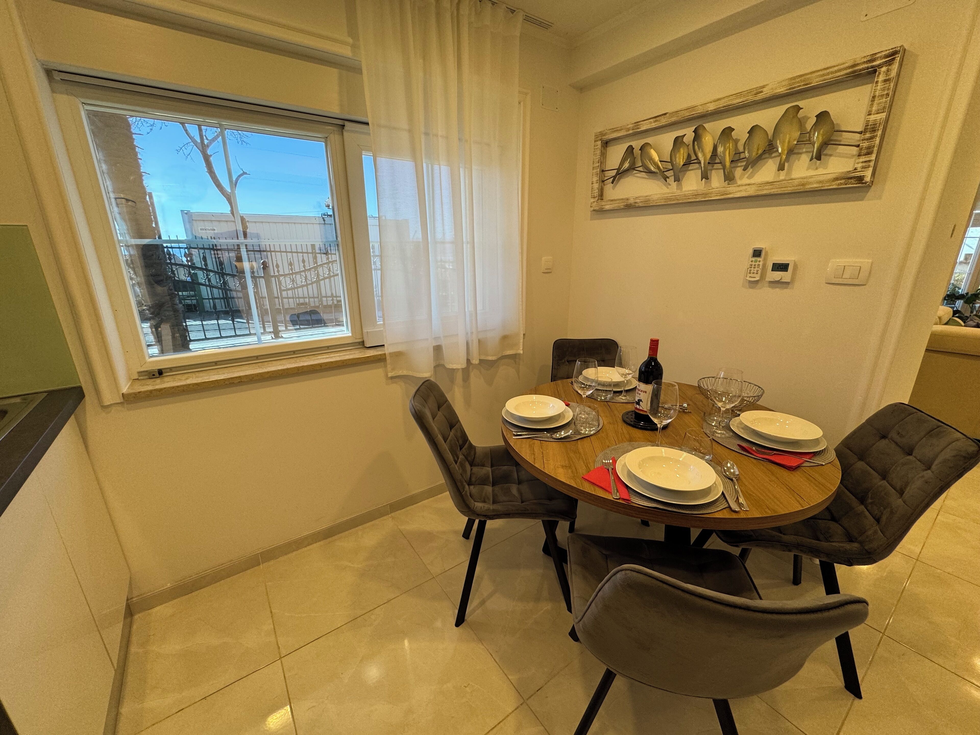 A dining area with a round table set for four, chairs, a wine bottle, and a wall-mounted bird art piece.