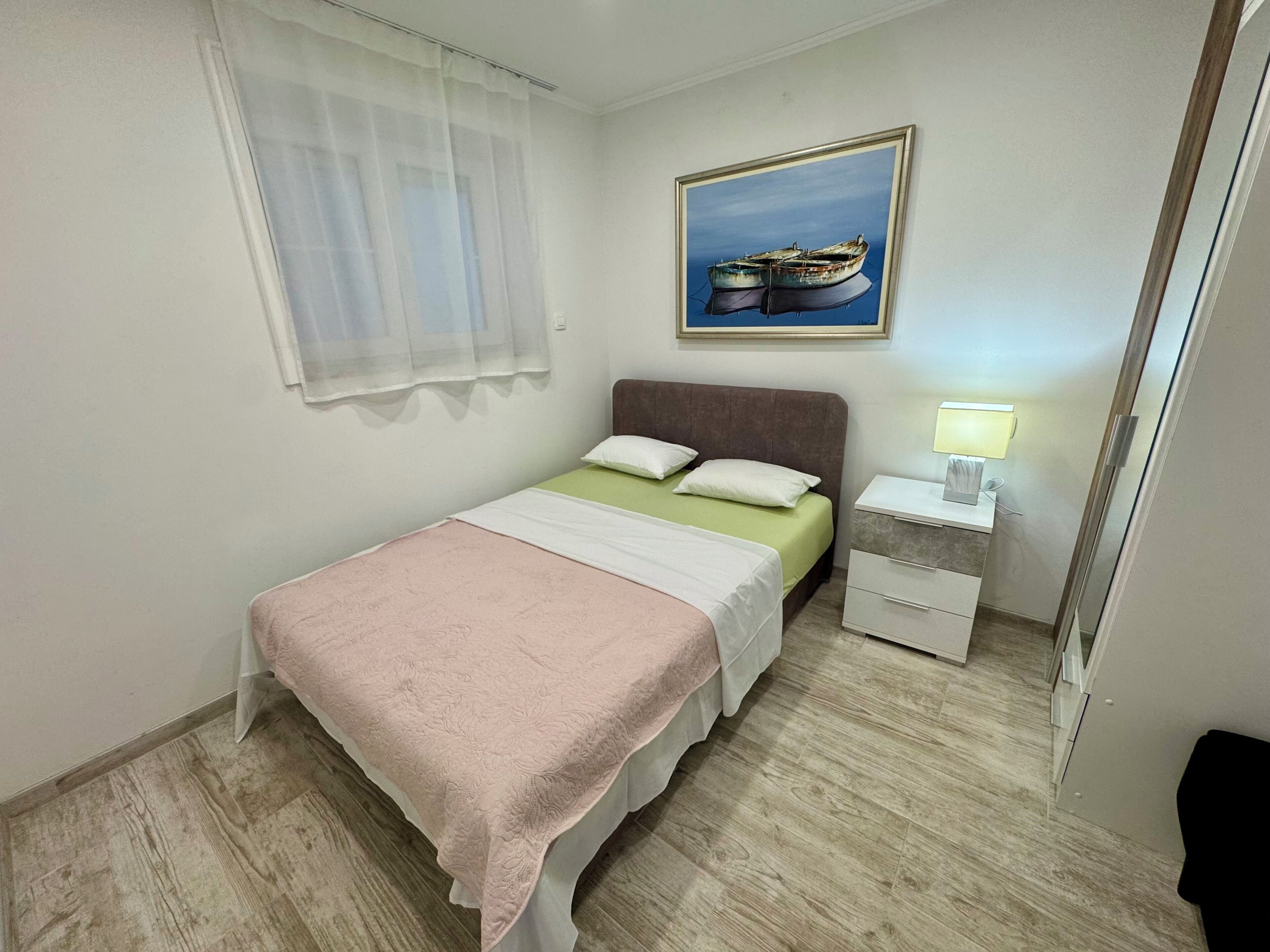Una camera da letto ordinata con un letto, un comodino con una lampada, un quadro appeso alla parete e una finestra con tende leggere.