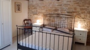 A bedroom with a stone wall, a metal bed frame, a wooden chair, and a bedside table with a lamp. - Villa (sleeps 6), private pool, gardens surrounded by countryside and woodland (Verfeil)