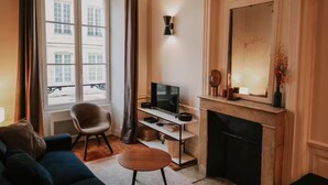 A living room with a sofa, a chair, a TV on a stand, a fireplace, and a large window with curtains. - Le Réaumur | Hyper Centre & Extérieur (La Rochelle)