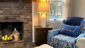 Ein gemütliches Wohnzimmer mit Kamin aus Backsteinen, einer Lampe und einem blau gemusterten Sessel mit einem Kissen mit dem Aufdruck "Awind".