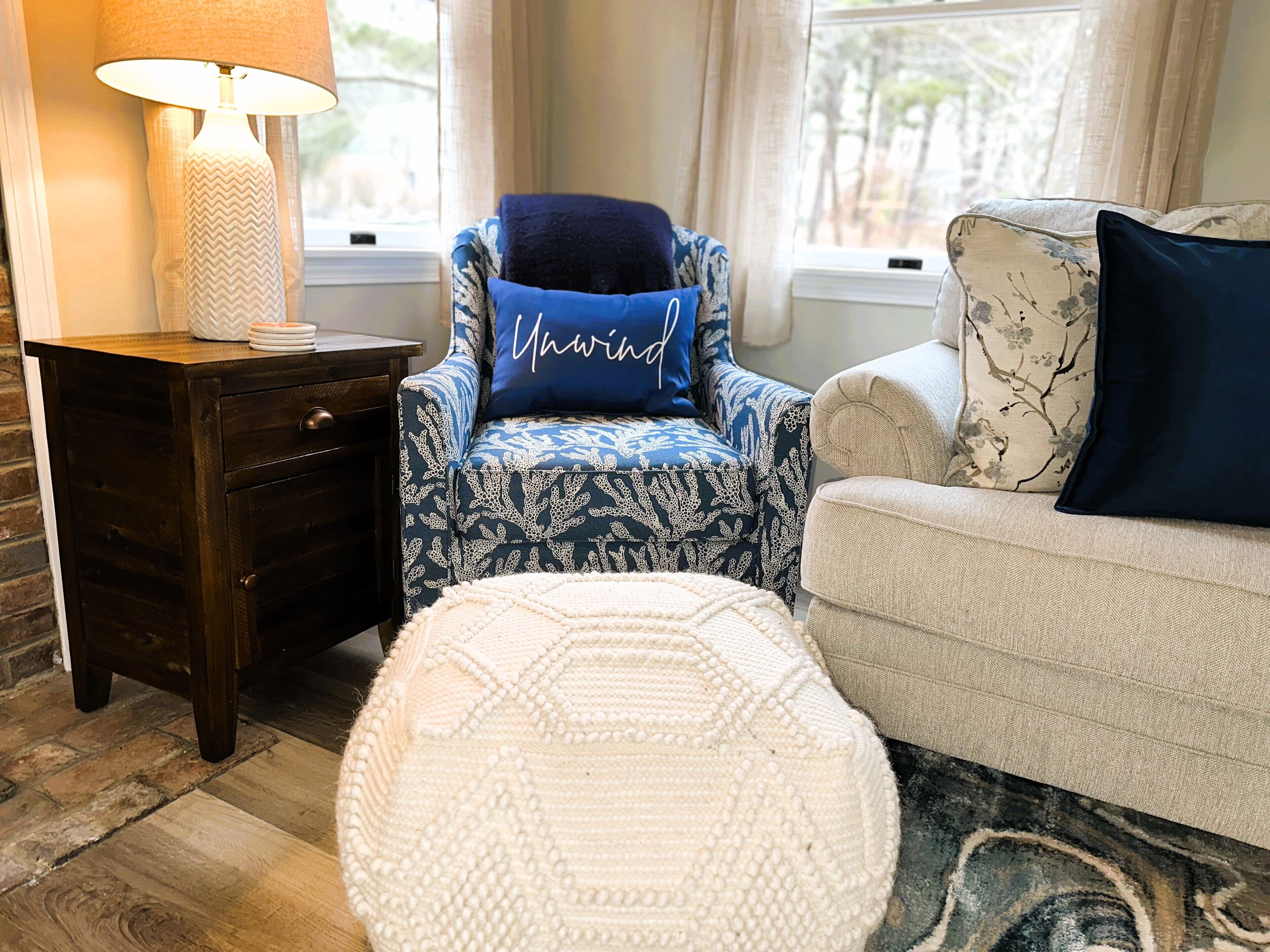 A cozy living room with a dark wooden side table, a blue patterned armchair with a 'Mawnd' pillow, a white ottoman, and a beige sofa with floral cushions.