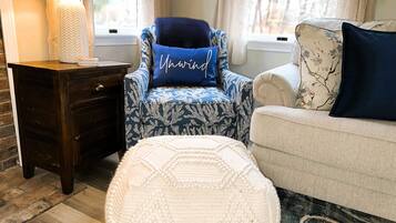A cozy living room with a dark wooden side table, a blue patterned armchair with a 'Mawnd' pillow, a white ottoman, and a beige sofa with floral cushions.