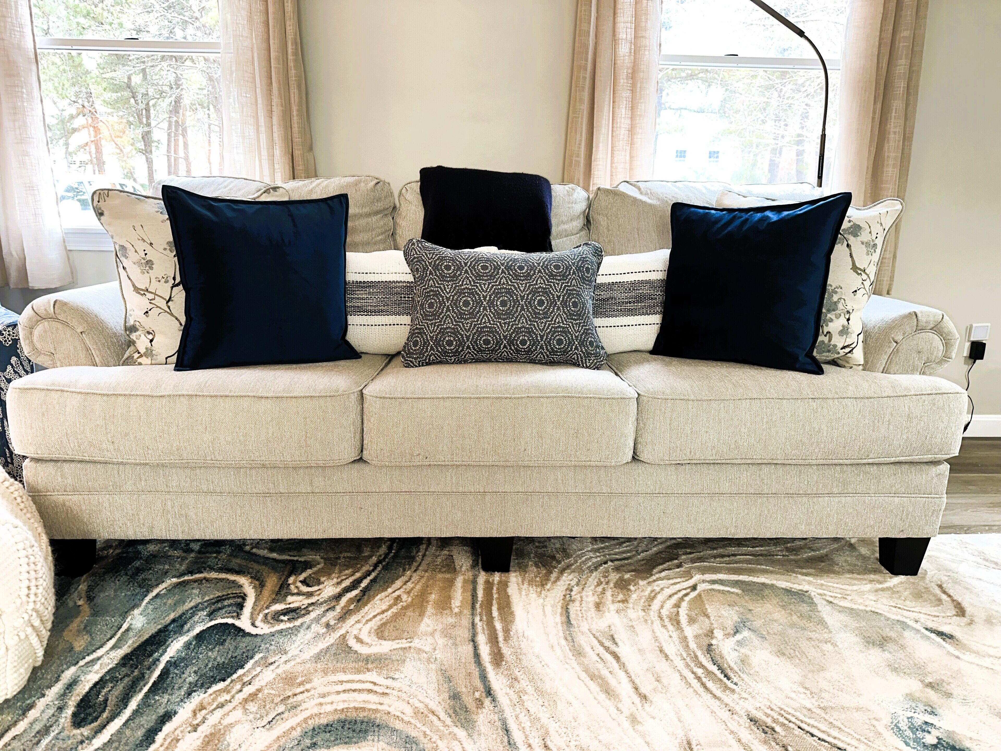 Un canapé beige agrémenté de coussins à motifs et un tapis à motifs de marbre dans une pièce dotée de grandes fenêtres et de rideaux.