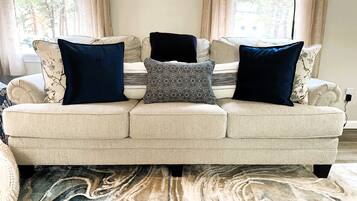 Un canapé beige agrémenté de coussins à motifs et un tapis à motifs de marbre dans une pièce dotée de grandes fenêtres et de rideaux.