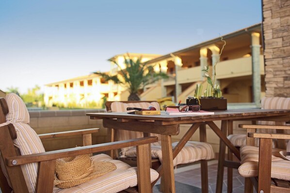 An outdoor seating area with striped cushioned chairs and a wooden table, featuring a book, glasses, and a potted plant.
