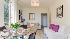 A modern living room with a glass dining table, beige sofa, and a white couch. There is a large window with curtains, a ceiling light, and a framed artwork on the wall. - BELLE DARSE AP4454 By Riviera Holiday Homes (Villefranche-sur-Mer)