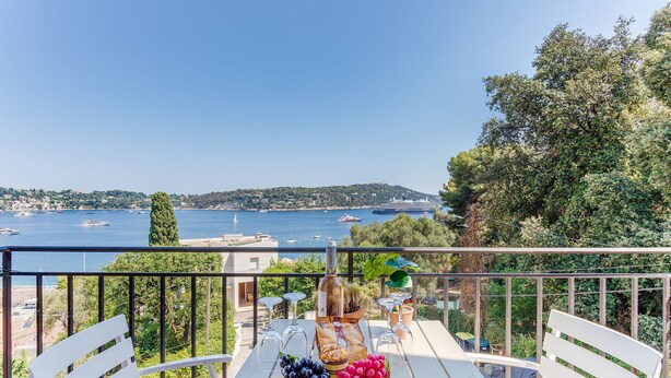A balcony with a table set for a meal, overlooking a scenic coastal view with boats and greenery. - BELLE DARSE AP4454 By Riviera Holiday Homes (Villefranche-sur-Mer)