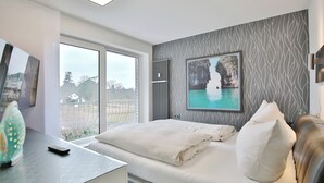 A modern bedroom with a large window, a bed with white linens, a painting of a mountain and lake, and a radiator. - Strandschlösschen Apartment 27 (Haffkrug)