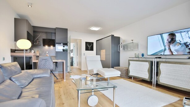 A modern living room with a grey sofa, a glass coffee table, a white armchair, a TV, and a kitchen area in the background. - Strandschlösschen Apartment 27 (Haffkrug)