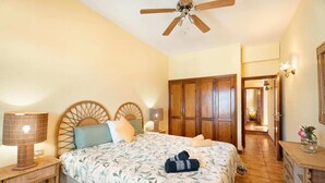 Une chambre à coucher comprenant un lit, une tête de lit en bois, des lampes de chevet, un ventilateur de plafond, une armoire et une petite table sur laquelle est posé un vase.