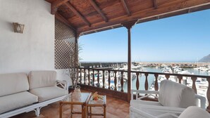 Un balcon avec un canapé blanc, des fauteuils en osier et une table garnie de fruits. Il offre une vue sur une marina avec des bateaux et un ciel bleu azur.