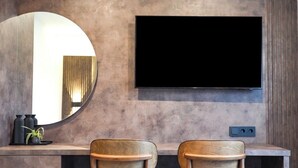 A modern room with a large flat-screen TV mounted on a wall, two wooden chairs, a round mirror, and a small plant on a shelf. - JG Hotel Antoniergasse (Memmingen)