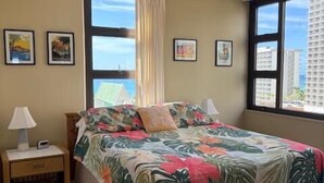 A bedroom with a bed, a nightstand, a lamp, and a window with a view of buildings. - Waikiki Banyan 1 Bedroom with Parking! (Honolulu)