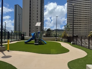A playground with a slide and climbing structure, surrounded by a well-maintained lawn and tall buildings in the background. - Waikiki Banyan 1 Bedroom with Parking! (Honolulu)