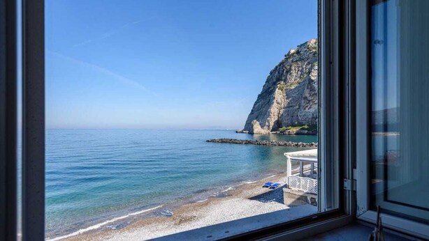 Vista costera desde una ventana con vista a una playa, un acantilado rocoso y un cielo despejado.