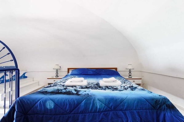 Ein Schlafzimmer mit gewölbter Decke, einem Bett mit blauer Bettdecke, zwei weißen Handtüchern und einem kleinen Nachttisch mit Lampe.