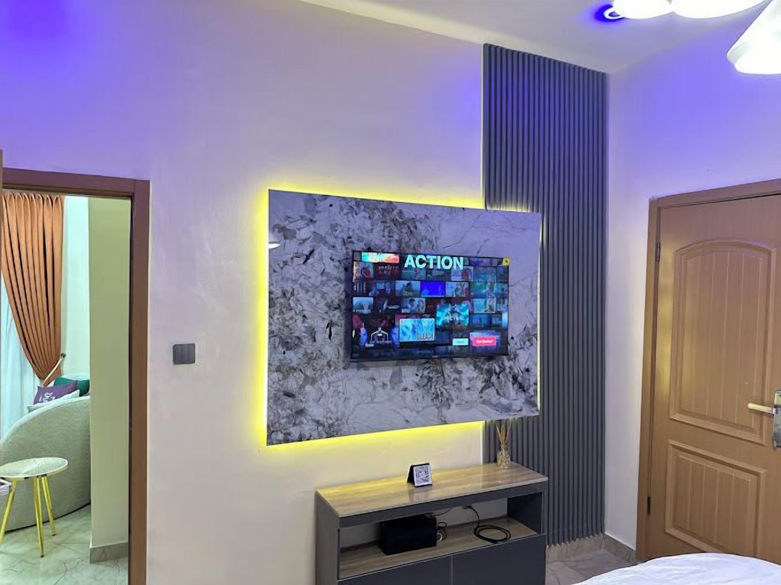 A modern bedroom with a large wall-mounted TV displaying various game options, a wooden bedside table, and a door with a vertical panel design.