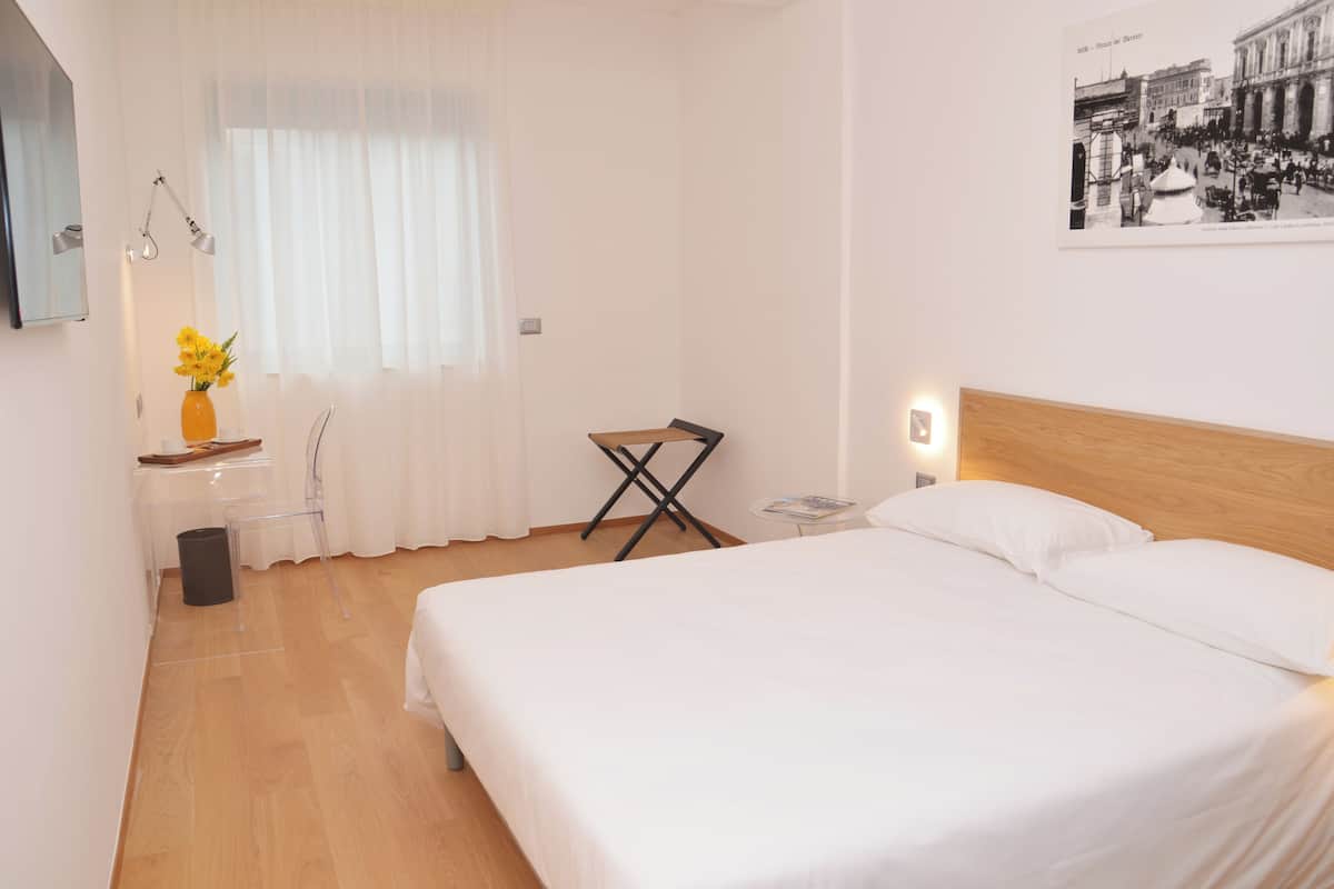 A hotel room with a bed, a desk with a chair, a small table with a lamp, and a picture on the wall.