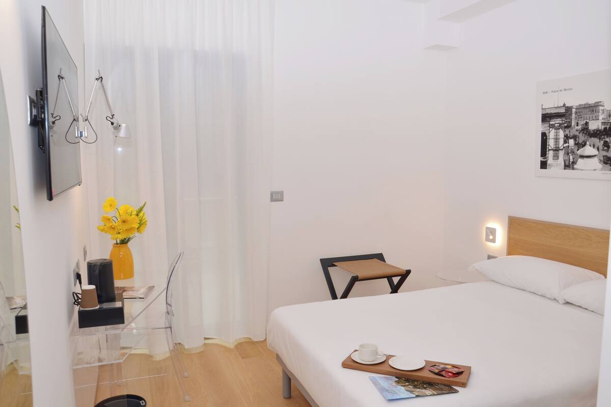 A hotel room with a bed, a desk with a chair, a small table with a tray, a vase with flowers, and a wall-mounted TV.