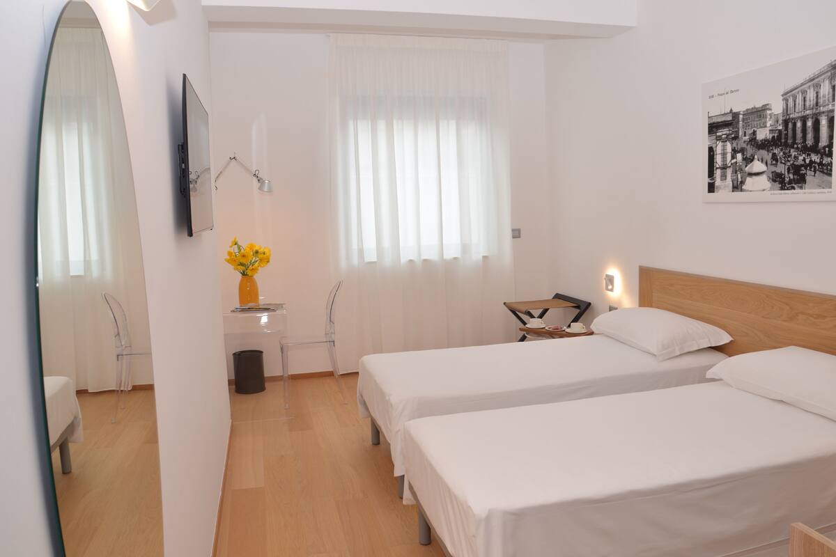 A hotel room with two single beds, a wooden headboard, a flat-screen TV mounted on the wall, a small table with a vase of flowers, and a window with sheer curtains.