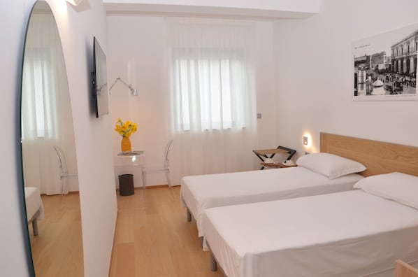 A hotel room with two single beds, a wooden headboard, a flat-screen TV mounted on the wall, a small table with a vase of flowers, and a window with sheer curtains.