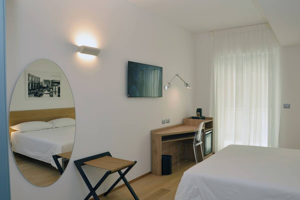 A hotel room with a bed, a desk, a chair, a mirror, a picture on the wall, and a window with curtains.