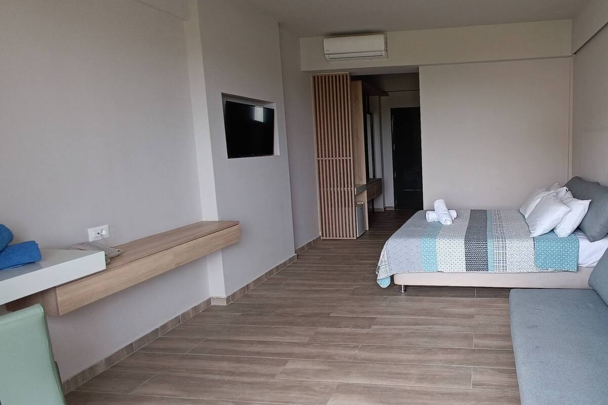 A modern hotel room with a sofa bed, a small desk with a chair, a flat-screen TV, and a wooden partition.
