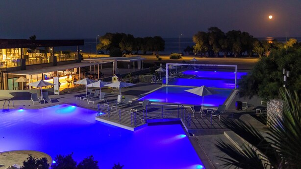 A nighttime pool area with blue lighting, lounge chairs, and a building with outdoor seating. - Gennadi Sea Side Resort (Rhodes)