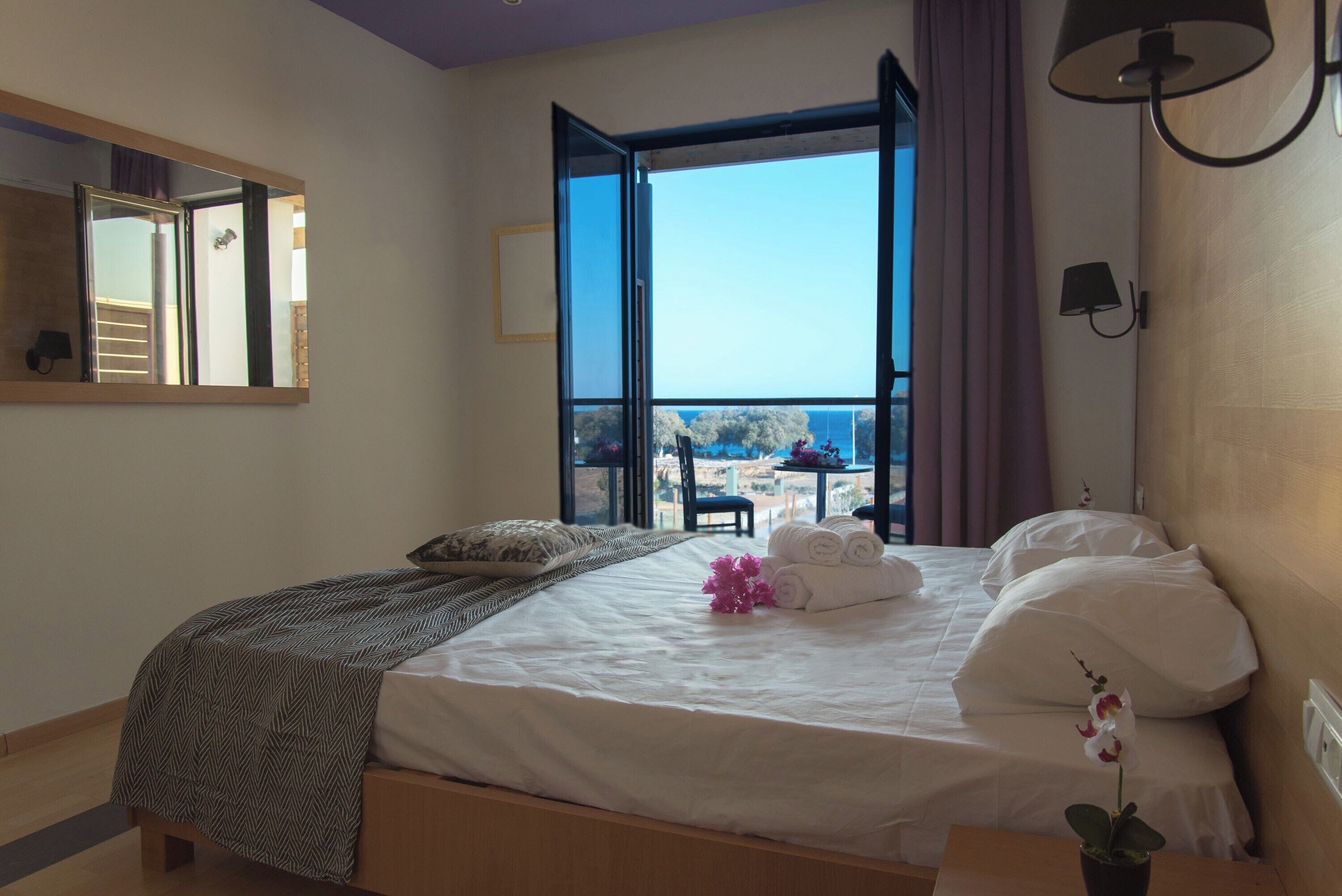 A bedroom with a large bed, a view of the sea, and a balcony with a table and chairs.