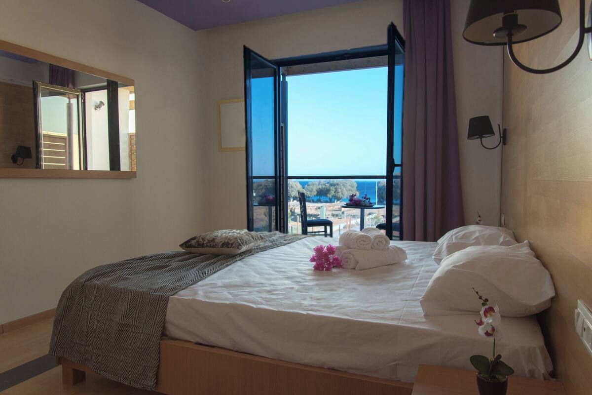 A bedroom with a large bed, a view of the sea, and a balcony with a table and chairs.