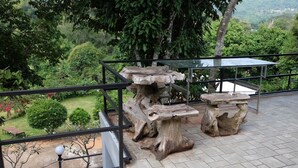 Een stenen tafel en bank staan op een betegeld terras met een metalen reling, met uitzicht op een weelderige tuin en een glooiend landschap.