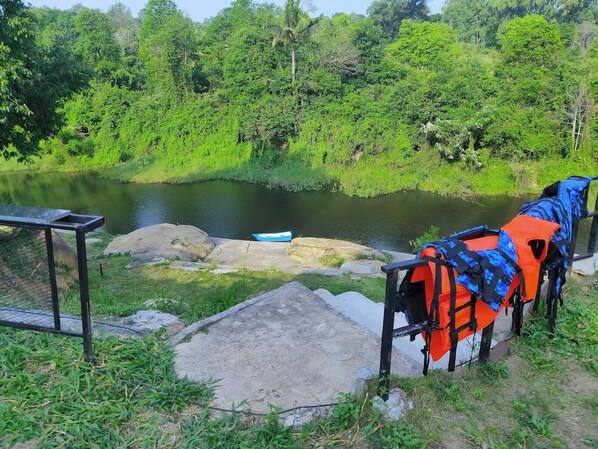 Un campamento con una mesa y sillas de hormigón, una canoa y un chaleco salvavidas sobre una roca cerca de un río tranquilo rodeado de vegetación densa.