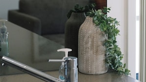 Un baño con un espejo, un lavamanos, un florero con una planta y un cuadro abstracto enmarcado en la pared.