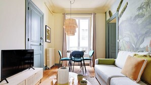 A living room with a television, a dining table with chairs, a sofa, and a large window with curtains.