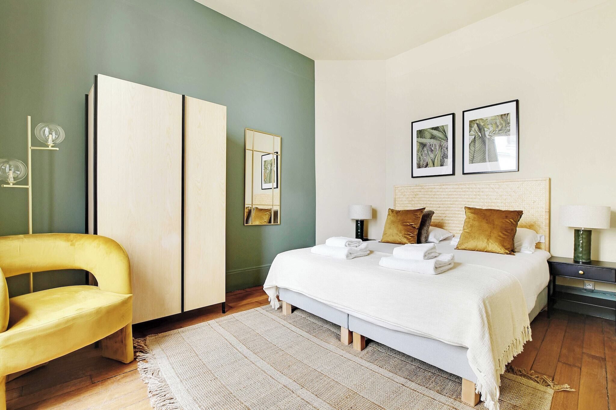 A bedroom with a bed, a yellow chair, a wardrobe, and two framed pictures on the wall.