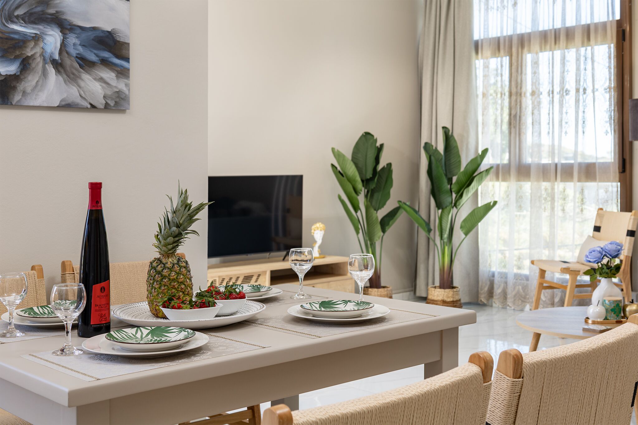 Un comedor con una mesa dispuesta para dos personas, con platos que presentan un diseño de hoja tropical, una botella de vino tinto y una piña.
