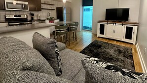 Una cocina moderna con una isla central, un comedor con taburetes de bar, una sala de estar con sofá y televisor, y un ventanal que proporciona luz natural.