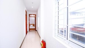 A corridor with a wooden door at the end, a window on the right, and a fire extinguisher on the floor. - Moonlight Quynhon Hotel (Quy Nhon Nam)
