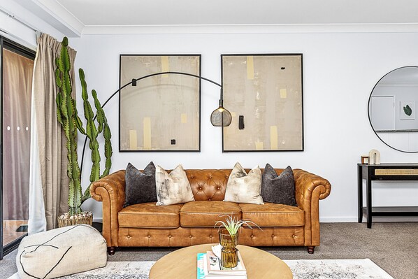 Un salon avec un canapé en cuir marron, deux tableaux abstraits, un cactus et une lampe sur pied.