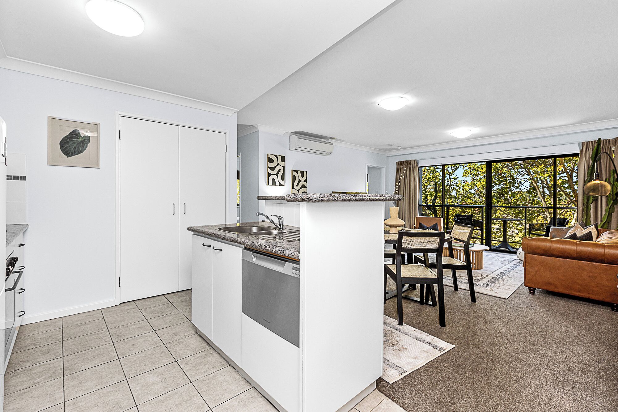 A modern kitchen with white cabinets and a granite countertop, a dining area with a brown leather sofa and a large window offering a view of trees.