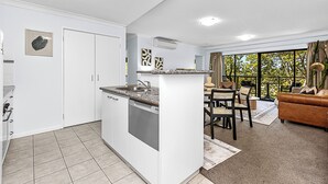 A modern kitchen with white cabinets and a granite countertop, a dining area with a brown leather sofa and a large window offering a view of trees. - Luxury Perth Getaway: Modern Design (Perth WA)