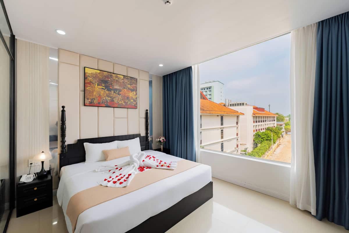 A hotel room with a large bed, a nightstand with a lamp, a painting on the wall, and a view of buildings through the window.