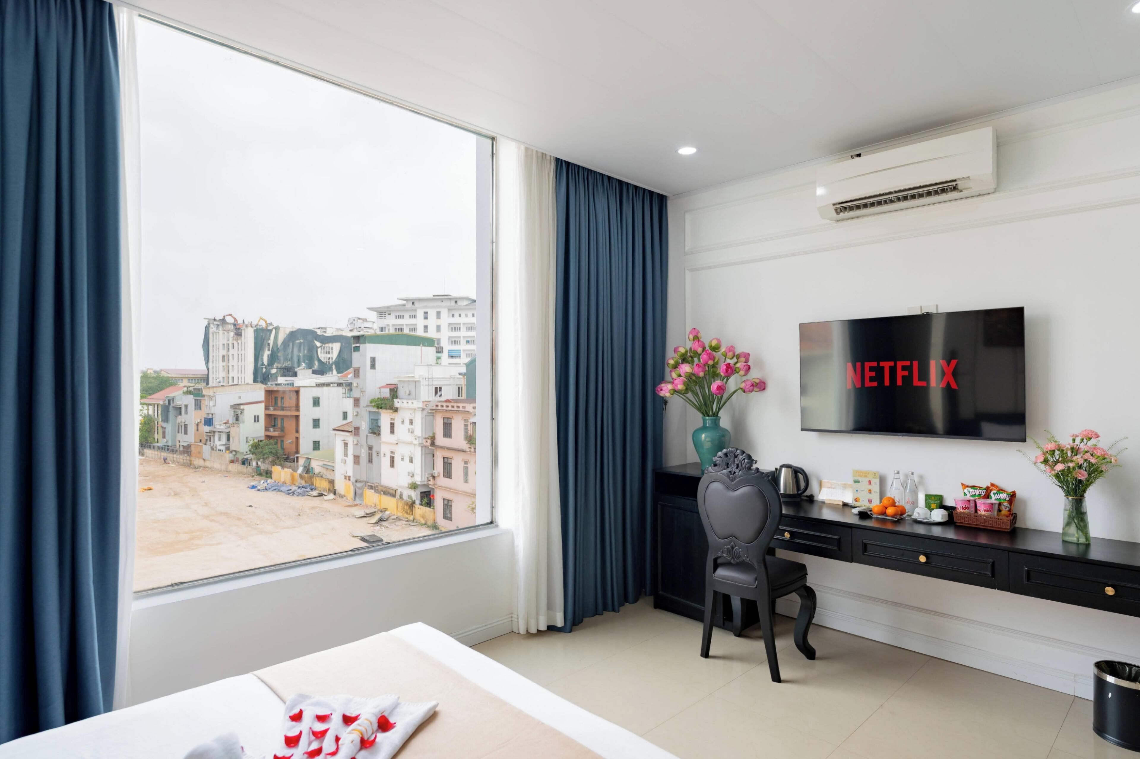 Ein Hotelzimmer mit großem Fenster, Schreibtisch mit Stuhl, Flachbildfernseher und einer Vase mit Blumen.