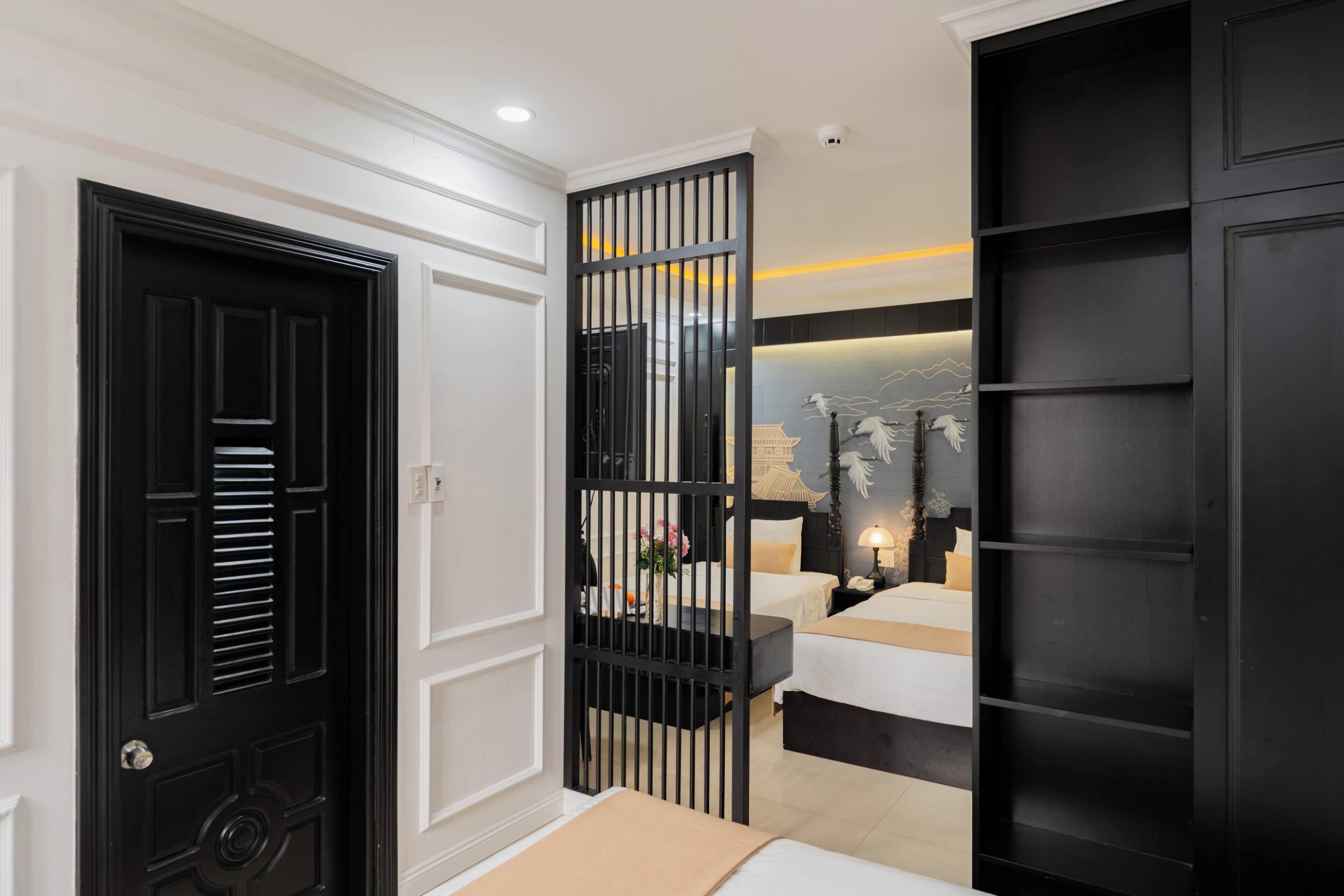A hotel room with a black door, a partition with vertical bars, a bed with a white headboard, a bedside table with a lamp, and a wall-mounted artwork.